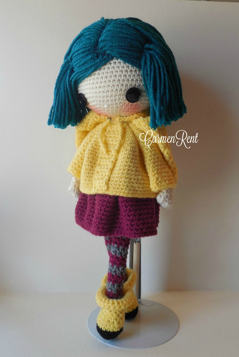 May include: Crocheted doll with teal hair, a yellow hooded sweater, a burgundy skirt, striped tights, and yellow boots. The doll is standing on a white display stand.