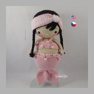 May include: A crocheted mermaid doll with pink and beige clothing, a white headband with pink stripes, and black hair. The doll is wearing a pink bikini top and a pink mermaid tail. The doll is standing on a clear stand.