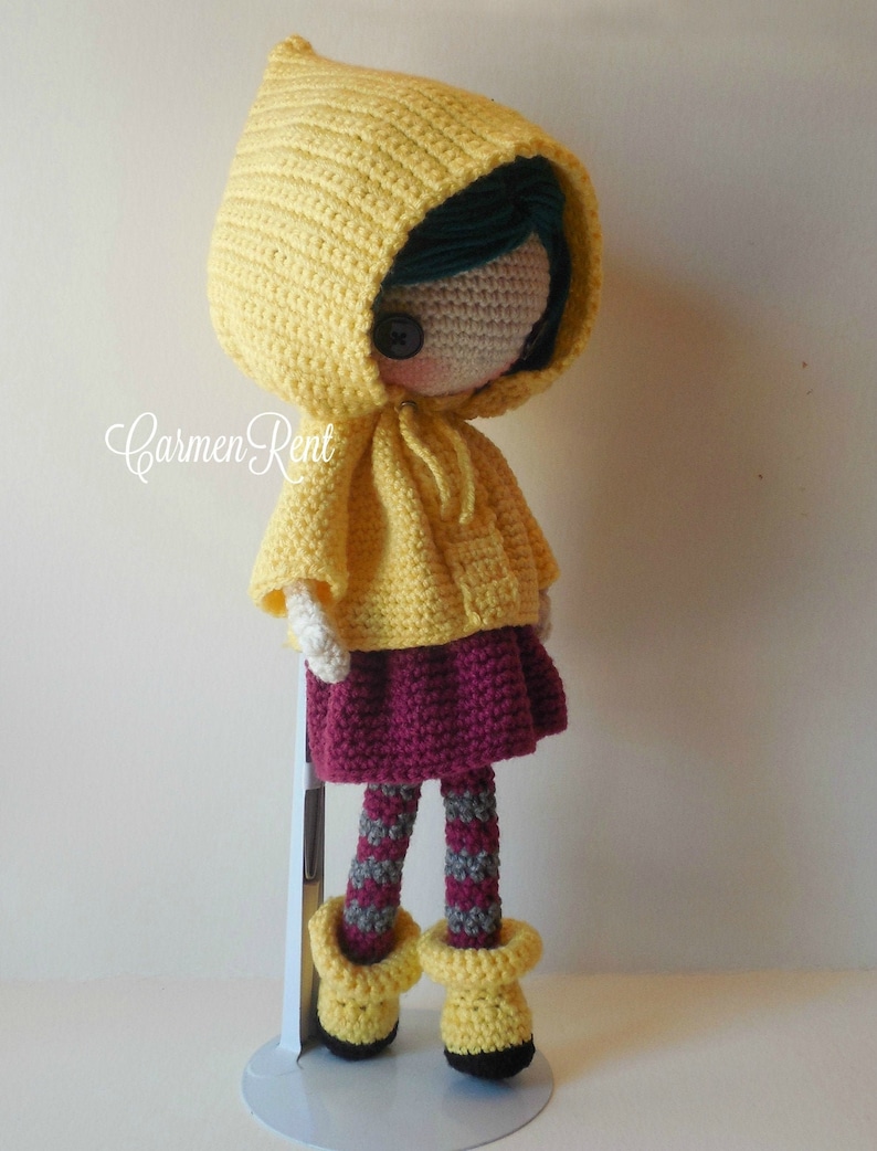 May include: A crocheted doll wearing a yellow raincoat with a hood, a burgundy skirt, and gray and white striped tights. The doll has a button nose and black eyes.