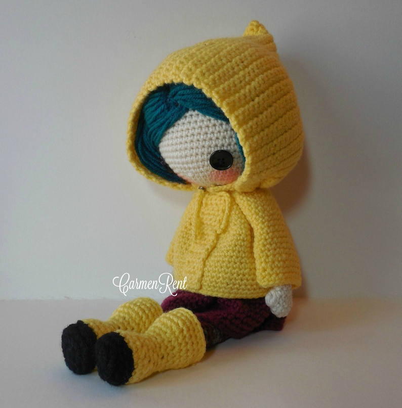 May include: A crocheted doll wearing a yellow raincoat with a hood, blue hair, and black boots. The doll is sitting on a white surface.