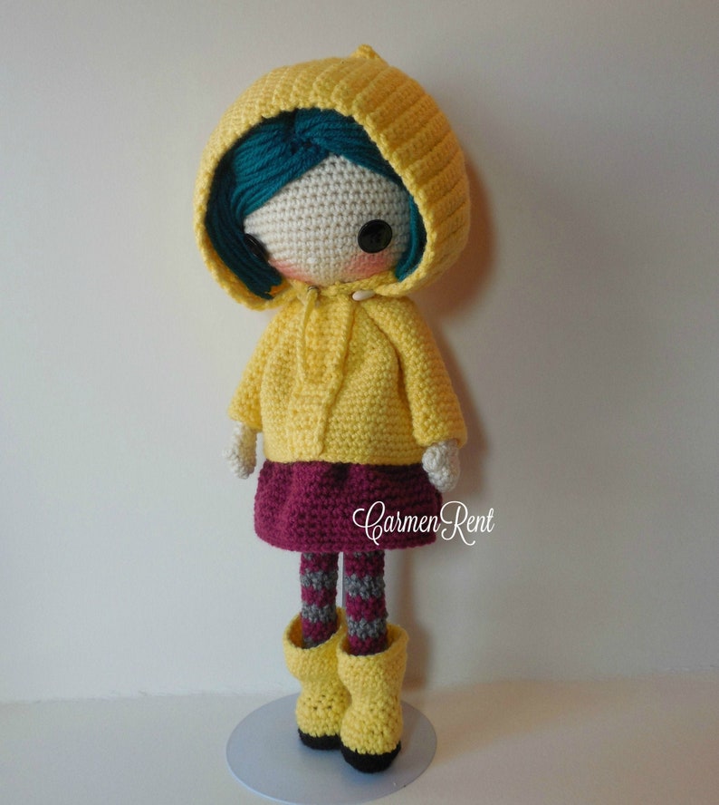 May include: A crocheted doll wearing a yellow raincoat, a burgundy skirt, and grey and white striped tights. The doll has teal hair and brown eyes. The doll is standing on a light blue base.