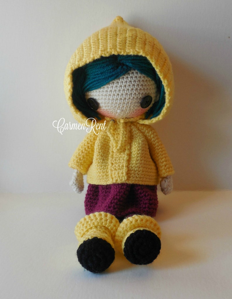 May include: A crocheted doll wearing a yellow raincoat with a hood, a burgundy skirt, and black boots. The doll has teal hair and black button eyes.