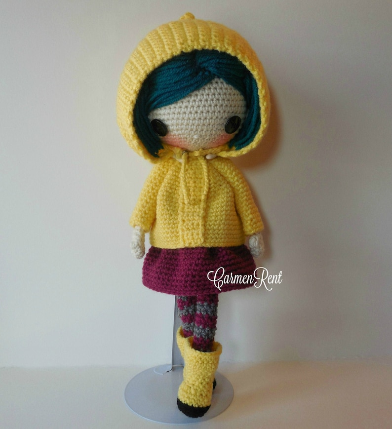 May include: Crocheted doll wearing a yellow raincoat, a burgundy skirt, and striped tights. The doll has teal hair and is standing on a white stand. The text "Carmen Rent" is visible on the doll's skirt.