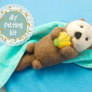 May include: A handmade felted otter figure, holding a yellow flower with green accents. The otter has brown fur, a white face, and black eyes. The image includes the text "DIY felting kit" in a decorative circle.