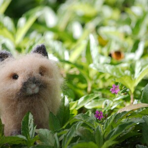 May include: A fluffy, light brown, felted animal with black eyes and a pink nose. The animal is sitting in a bed of green leaves.