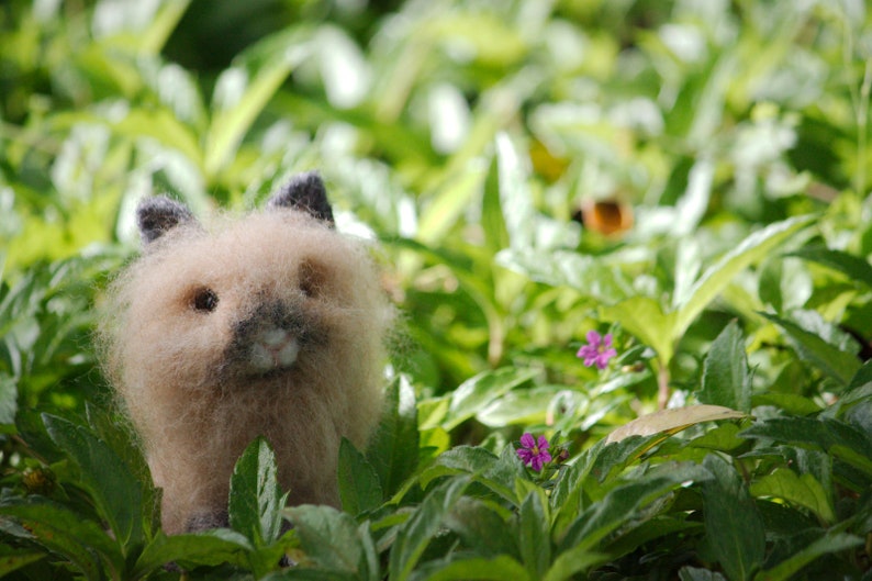 May include: A fluffy, light brown, felted animal with black eyes and a pink nose. The animal is sitting in a bed of green leaves.
