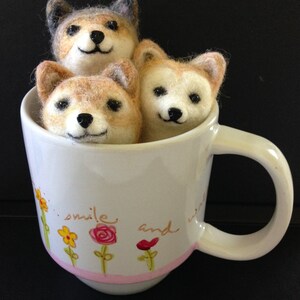 May include: Three felted dog figurines, one grey and two brown, are sitting inside a white ceramic mug with a pink rim. The mug has a floral design and the text "smile and" printed on the side.
