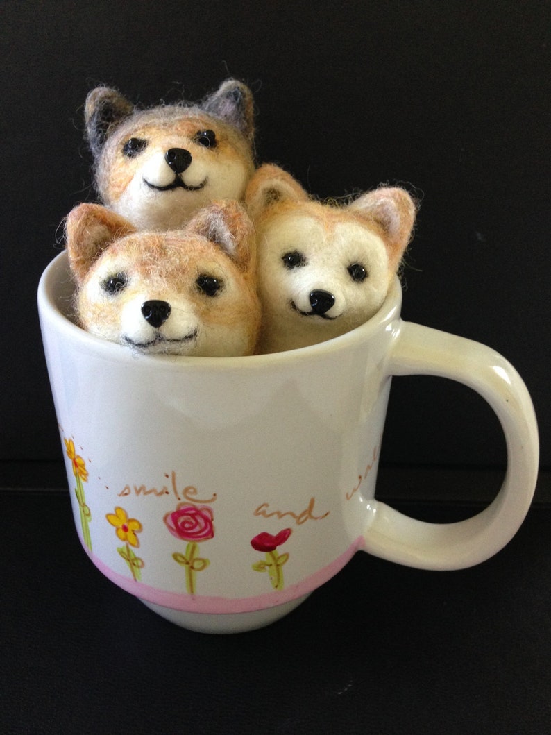 May include: Three felted dog figurines, one grey and two brown, are sitting inside a white ceramic mug with a pink rim. The mug has a floral design and the text "smile and" printed on the side.