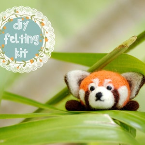 May include: DIY felting kit featuring a red panda with black ears and legs. The red panda is sitting in a bed of green grass.