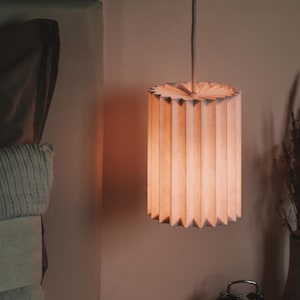 May include: A cylindrical paper lampshade hangs above a nightstand. The lampshade is made of vertical, folded paper strips. A vintage alarm clock, an open book, and a vase with dried plants sit on the nightstand.