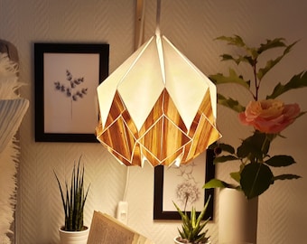 Puede incluir: Una mesita de noche con un libro, una vela y dos plantas en maceta. Una lámpara de estilo origami de madera y blanca cuelga sobre la mesa.