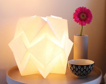 Könnte beinhalten: Eine weiße Lampe im Origami-Stil mit warmem Schein steht auf einem weißen Beistelltisch neben einem gelben Sessel mit einem weißen, flauschigen Kissen. Eine rosa Blume in einer grauen Vase steht ebenfalls auf dem Tisch.
