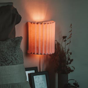 Könnte beinhalten: Ein pfirsichfarbener, plissierter Lampenschirm ist an einer Wand montiert und wirft einen warmen Schein. Die Lampe ist rechteckig und hat ein vertikales, ziehharmonikaartiges Design. Ein Kissen, gerahmte Bilder, eine Vase mit getrockneten Blumen und ein Wecker sind ebenfalls zu sehen.