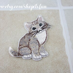 May include: Embroidered patch of a gray and white kitten sitting upright. The cat has blue eyes, a white chest, and brown accents. The patch is on a light beige surface. The text "www.etsy.com/shop/lxfun" is at the top.