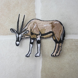 May include: Embroidered patch featuring an oryx, a large antelope with long, straight horns. The patch has a tan body with black and white markings on the legs and face. The horns are black, and the patch is likely for clothing or accessories.