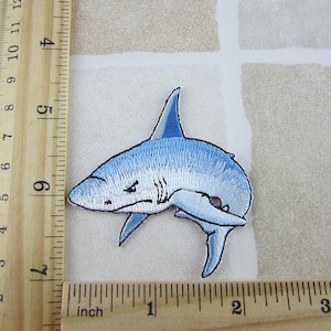 May include: Embroidered patch featuring a light blue shark with a white belly and a dark blue fin. The shark has an angry expression. The patch measures approximately 3 inches in length.