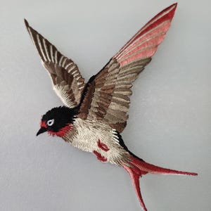 May include: Embroidered bird patch in flight, featuring detailed stitching. The bird has a black head, red accents, and a mix of brown, beige, and red feathers. The wings are spread wide, showcasing the intricate design.