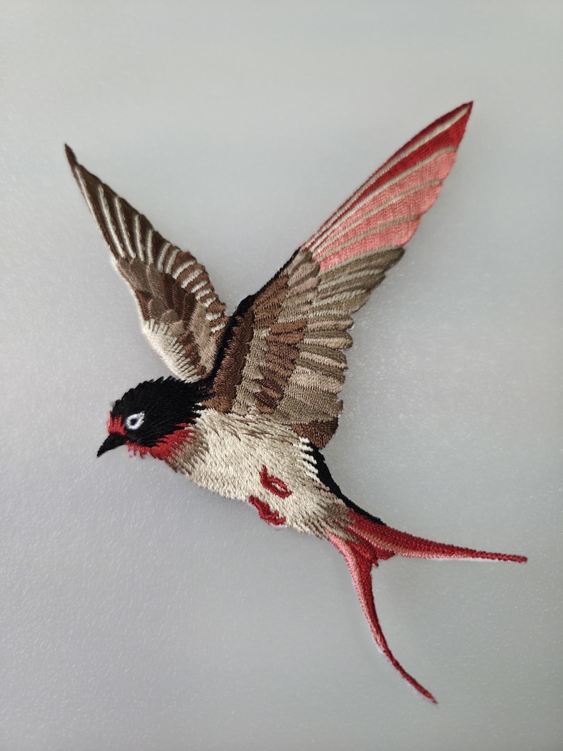 May include: Embroidered bird patch in flight, featuring detailed stitching. The bird has a black head, red accents, and a mix of brown, beige, and red feathers. The wings are spread wide, showcasing the intricate design.