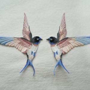 May include: Two embroidered swallow patches featuring black heads, white chests, and wings with shades of dusty pink and light blue. The birds are depicted in flight with wings spread wide, showcasing detailed stitching and a pointed tail design.