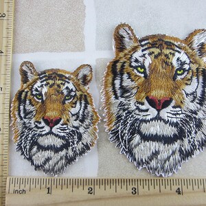 May include: Two embroidered tiger head patches. The larger patch is approximately 4 inches wide. The patches feature detailed stitching in shades of brown, black, and white, with green eyes and a red nose. The patches are on a neutral background.