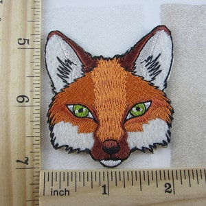 May include: Embroidered fox face patch with orange, white, and brown details. The fox has green eyes and a black outline. The patch measures approximately 7.6 cm wide and 7.6 cm tall.