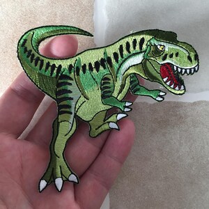 Può includere: Toppa ricamata di un dinosauro Tyrannosaurus Rex verde con strisce nere e bocca rossa. Il dinosauro ha artigli bianchi e un occhio giallo. La toppa è probabilmente per abbigliamento o accessori.