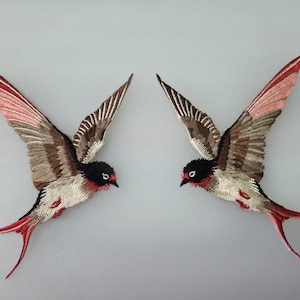Może przedstawiać: Dwa haftowane jaskółki z rozłożonymi skrzydłami. Ptaki mają czarne głowy, czerwone akcenty i skrzydła w odcieniach brązu, szarości i różu. Ich długie pióra ogonowe są czerwone. Jaskółki są wyświetlane na jasnoszarym tle.