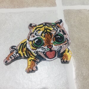 Puede incluir: Parche bordado de un cachorro de tigre de dibujos animados con grandes ojos verdes, una boca roja abierta y un patrón de rayas amarillas, negras y marrones. El parche está diseñado para coser o planchar sobre tela.