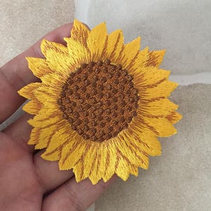 May include: Embroidered sunflower patch with yellow petals and a brown center. The patch is a detailed representation of a sunflower, with intricate stitching creating texture and depth. The bright colors and floral design make it suitable for various craft projects.