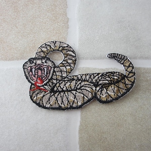 May include: Embroidered patch featuring a coiled snake with an open mouth, revealing a red tongue. The snake has a detailed pattern in shades of brown, tan, and black, with a black outline. The patch is designed to be sewn onto fabric.