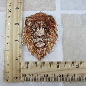Puede incluir: Parche bordado con la cabeza de un león, con melena marrón y ojos amarillos. El parche mide aproximadamente 9 cm de alto y 6,5 cm de ancho. El parche está sobre un fondo blanco.