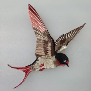 May include: Embroidered bird patch in flight, featuring a detailed design. The bird has a black head, red accents, and wings with shades of brown, beige, and red. The tail is red, and the patch is on a light gray background.