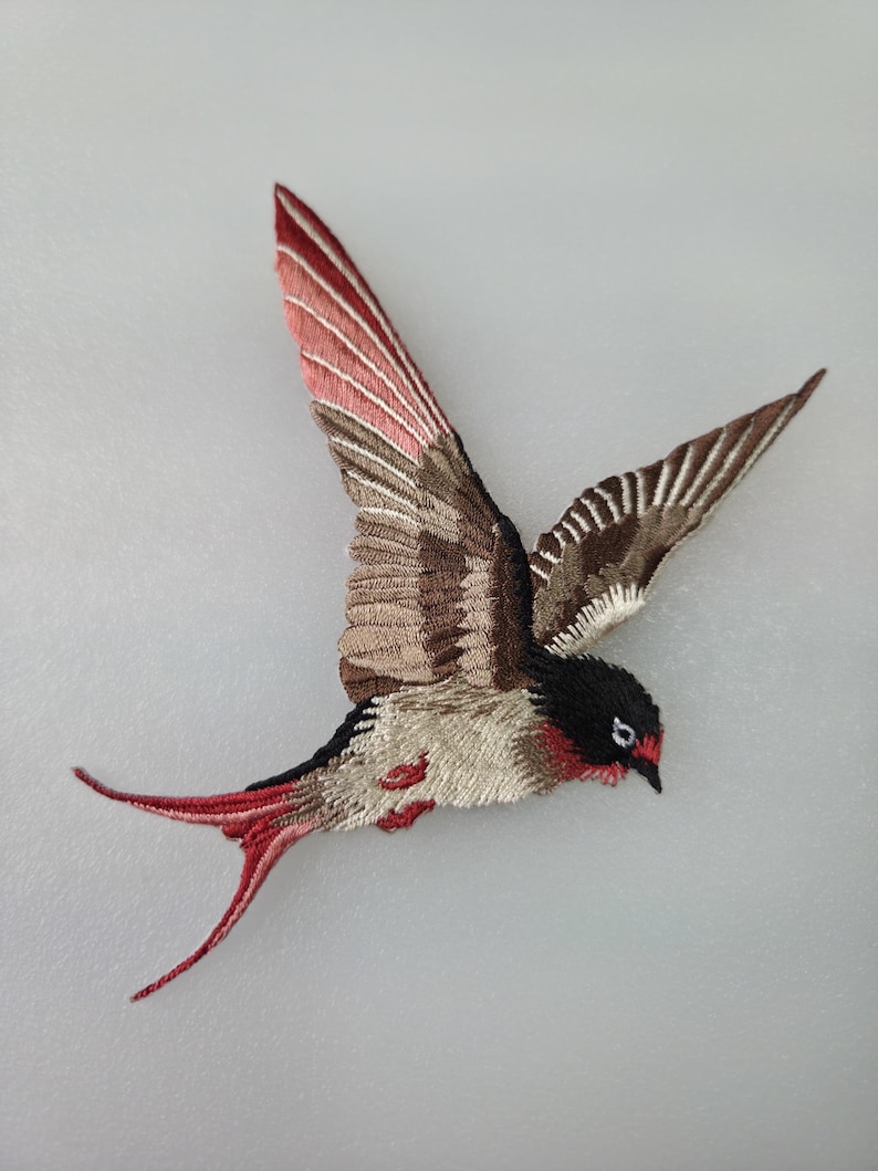 May include: Embroidered bird patch in flight, featuring a detailed design. The bird has a black head, red accents, and wings with shades of brown, beige, and red. The tail is red, and the patch is on a light gray background.