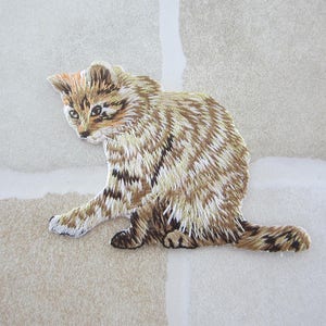 May include: Embroidered patch of a small cat in shades of brown, tan, and cream. The cat is depicted in a seated position, with its front paw extended. The patch has detailed stitching, creating a textured appearance. The cat's fur has a striped pattern.