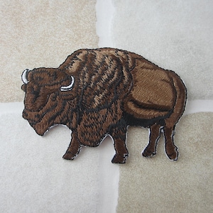 Op de afbeelding: Geborduurde patch met een bruine bizon, gedetailleerde vacht en witte hoorns. De patch toont de bizon in een wandelhouding. Ideaal voor het personaliseren van jassen, tassen of andere stoffen items. Ongeveer 10 cm lang.