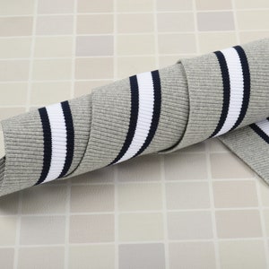 May include: Rolled gray ribbed knit fabric with a white stripe and a navy blue stripe. The fabric is a soft, flexible material, suitable for various sewing projects. The fabric is rolled up, showing the texture and design.