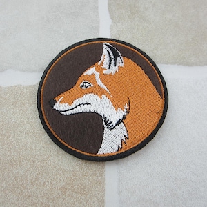May include: Embroidered patch with a fox profile in orange, white, and brown. The fox is set against a dark brown background within a circular shape, with a black border. Ideal for customising clothing or accessories.