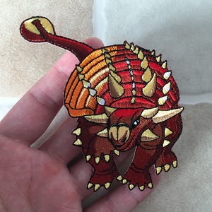 May include: Embroidered patch depicting an Ankylosaurus dinosaur. The patch uses a colour scheme of red, orange, and gold, with detailed stitching outlining the dinosaur's armoured body and spiky tail. The design is a detailed representation of the prehistoric creature.