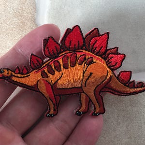 May include: Embroidered patch featuring a Stegosaurus dinosaur. The patch has a detailed design with orange and red colors. The dinosaur's plates and body are intricately stitched, creating a textured appearance. This patch is suitable for clothing or accessories.