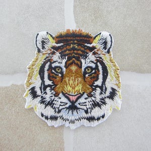 May include: Embroidered tiger head patch. The patch is intricately designed with brown, black, yellow, and white thread. The tiger's face features a pink nose and yellow eyes, with black stripes and white fur.