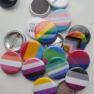 May include: A collection of circular pin-back buttons featuring various pride flag designs, including horizontal stripes in rainbow, transgender, bisexual, lesbian, asexual, and non-binary patterns, alongside white buttons with text and small heart icons.