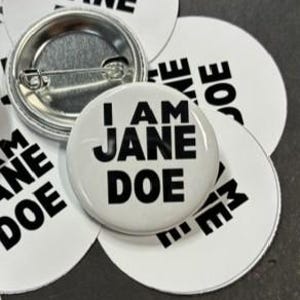 May include: A collection of circular white pin-back buttons featuring the black text I AM JANE DOE printed in a bold sans-serif font. One button is flipped over to reveal the silver-toned metal backing and the attached horizontal safety pin mechanism.