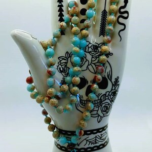 May include: A beaded necklace with turquoise and tan beads is draped over a white ceramic hand. The hand is decorated with black line art, including a skull, flowers, and geometric shapes. The necklace has a brown and turquoise tassel.