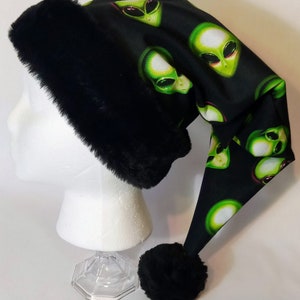 May include: A black Santa hat with a black faux fur trim and a black pom-pom. The hat features a repeating pattern of green alien faces on a black background. The hat is displayed on a white mannequin head.