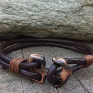 May include: A brown leather bracelet with a copper anchor clasp. The bracelet is double-layered and has a brown leather strap.
