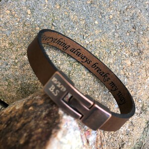 K&ouml;nnte beinhalten: Braunes Lederarmband mit einem kupfernen Verschluss. Das Armband ist mit dem Text "Everything always breaks my way" und dem Datum "12/19" sowie den Initialen "BS" graviert.