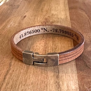 May include: A light brown leather bracelet with a rectangular metal clasp. The bracelet has three parallel lines etched into the leather and features the engraved coordinates 41.876500 °N, -72.703055 °W.