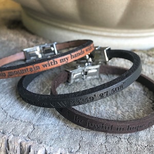May include: Three leather bracelets with different engraved messages. The top bracelet is brown and reads "I climb this mountain with my hands wide". The middle bracelet is black and reads "129W W 38°59' 27.508"". The bottom bracelet is brown and reads "Be joyful in hope."