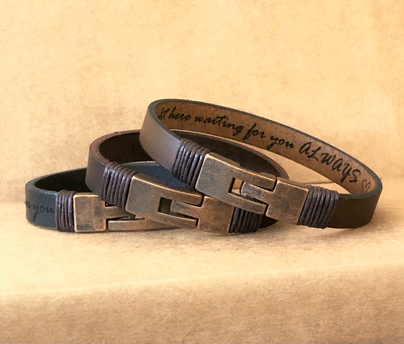 May include: Three leather bracelets in varying shades of brown and black, featuring a metal clasp and decorative stitching. One bracelet has the inscription "here waiting for you always". The bracelets are a stylish accessory.