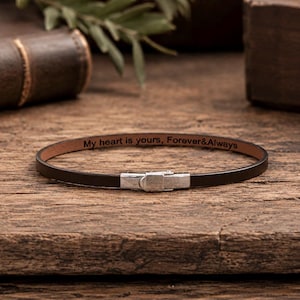 May include: A thin, dark brown leather bracelet featuring a silver-toned metal clasp. The interior surface of the leather band is engraved with the black text, My heart is yours, Forever&Always, centered along the length of the bracelet.